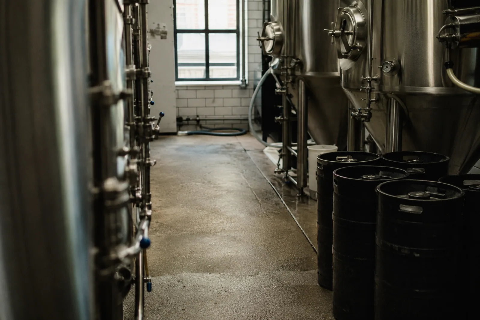 A small independent brewery with stainless fermenting tanks and unbranded dark kegs on worn concrete, available daylight and white tile, no people.