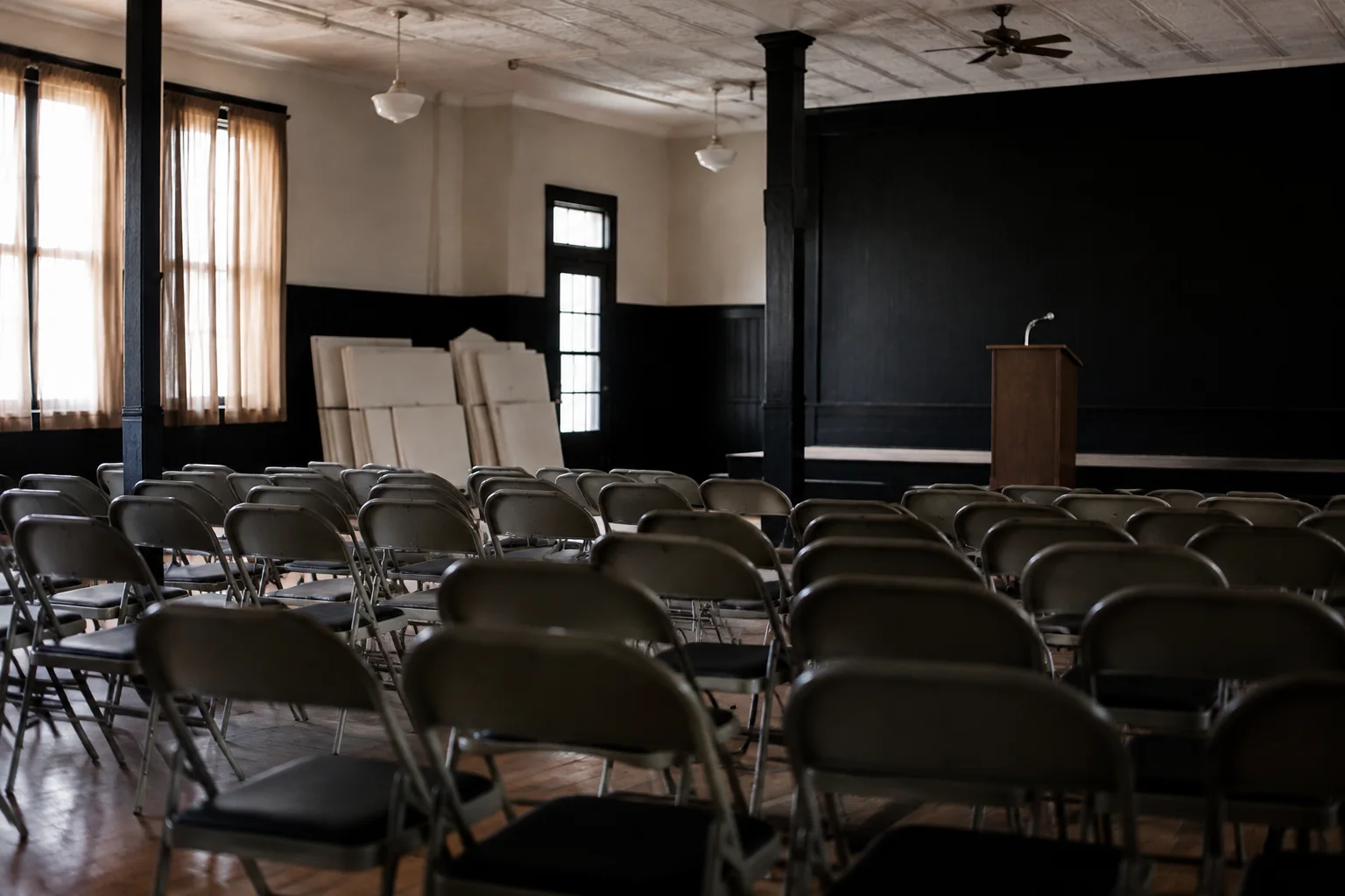 A local campaign rally venue just before opening, rows of empty folding chairs facing a small simple lectern in a community hall, blank placards stacked along a wall, no flags or real candidate identity.