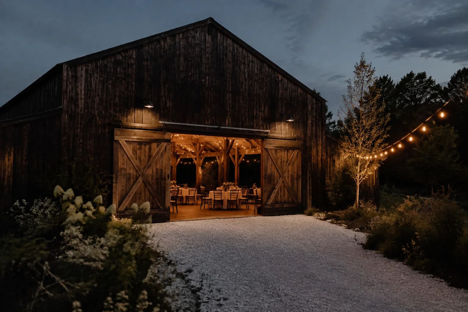 An events barn exterior at dusk, weathered dark timber, wide open doors revealing softly lit tables, white gravel path and simple string lights, no people.