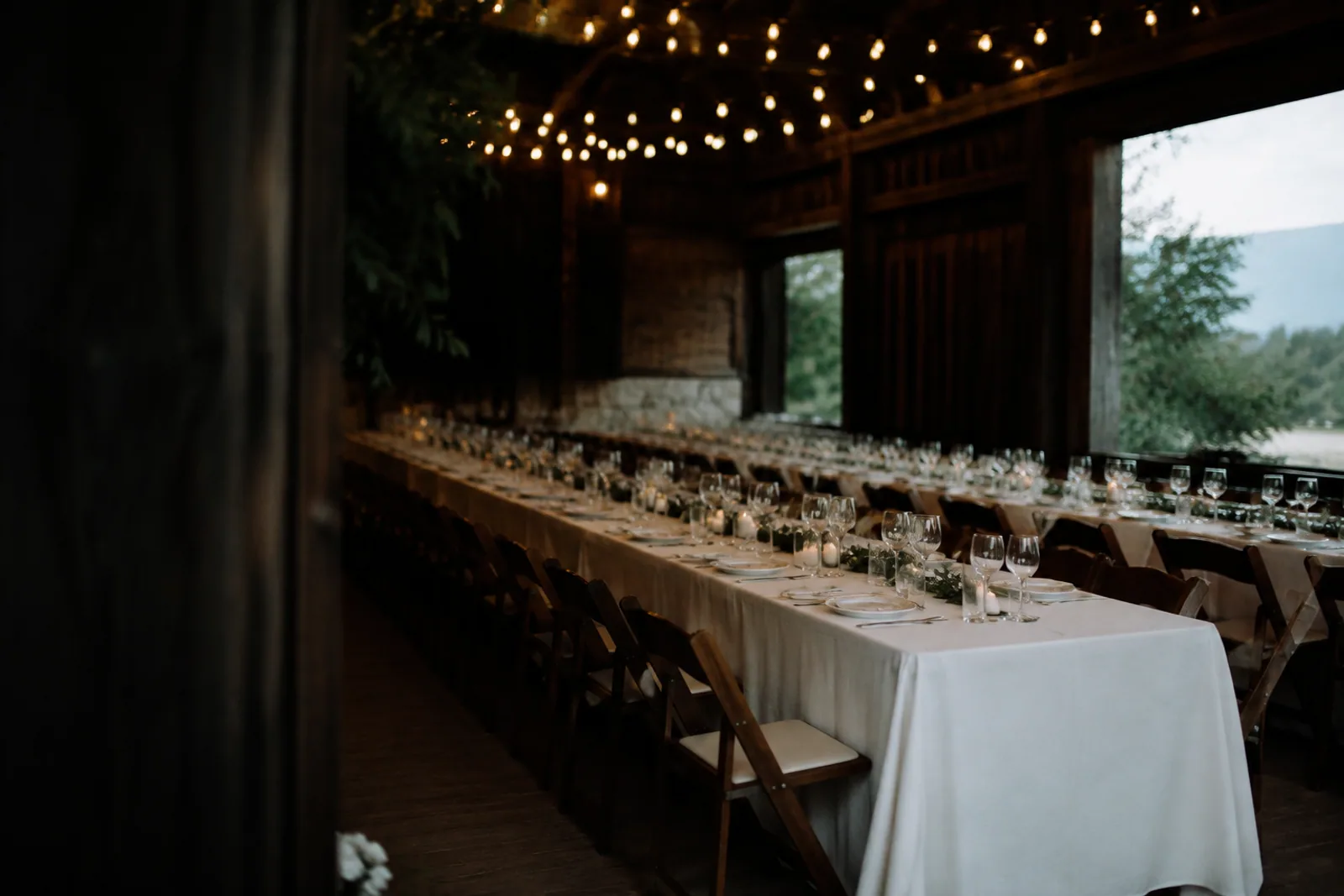 String lights above a long barn table set with white linen and simple glassware before an event, dark timber and dusk light, no people.