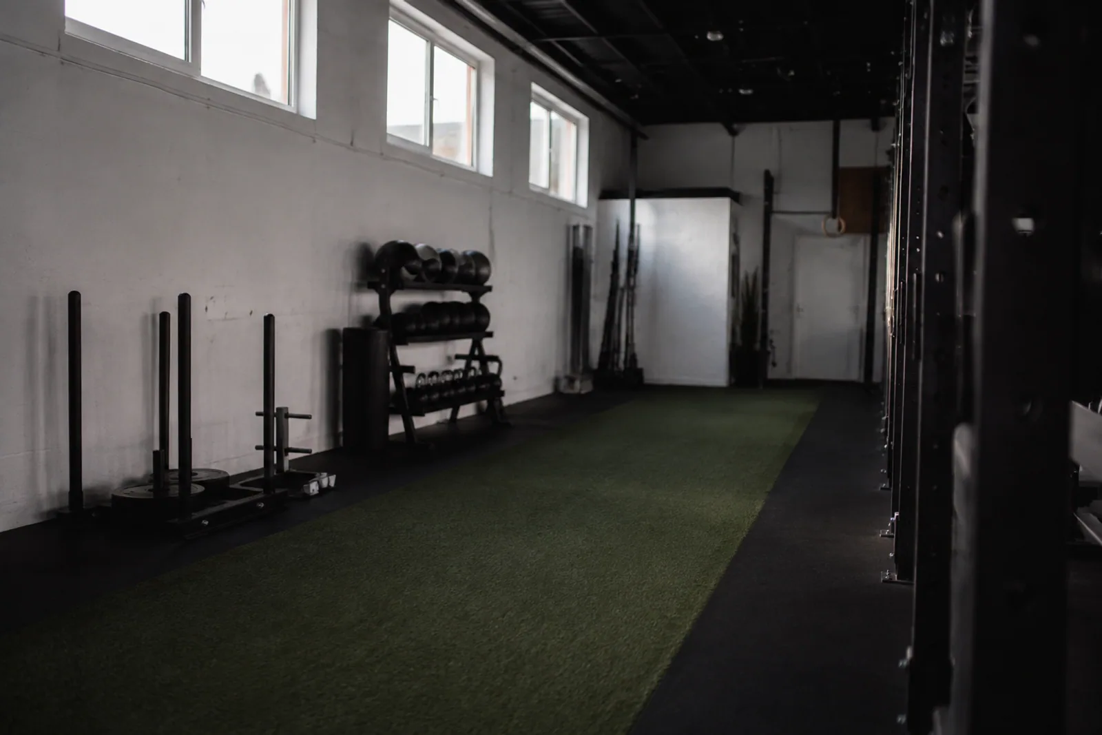 An empty narrow turf training lane at dawn in a neighborhood gym, charcoal racks and white walls, cool natural side light, no people.