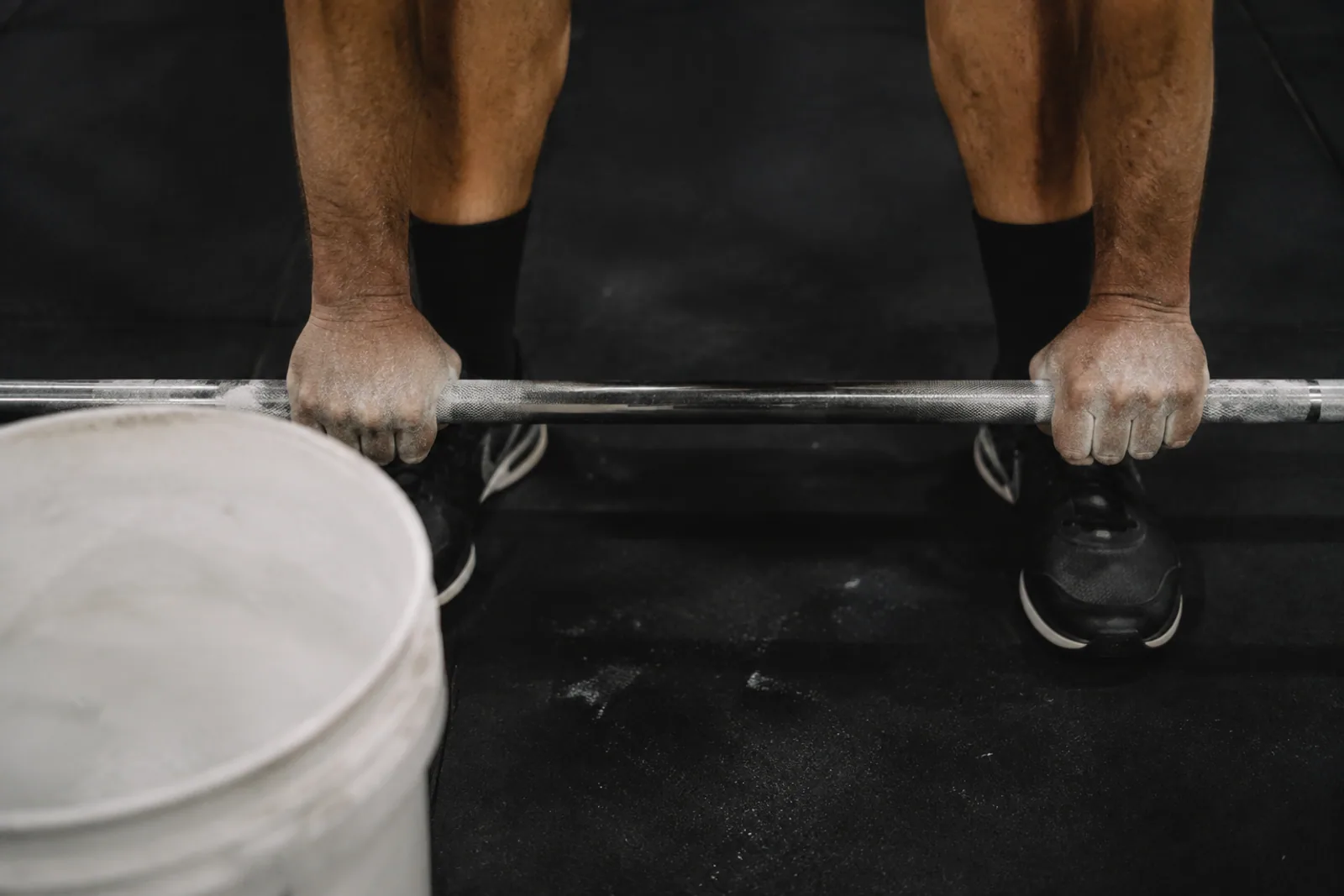 Cropped adult chalked hands gripping a steel barbell, black rubber floor and a white chalk bucket; natural hand anatomy, no face, no branding.