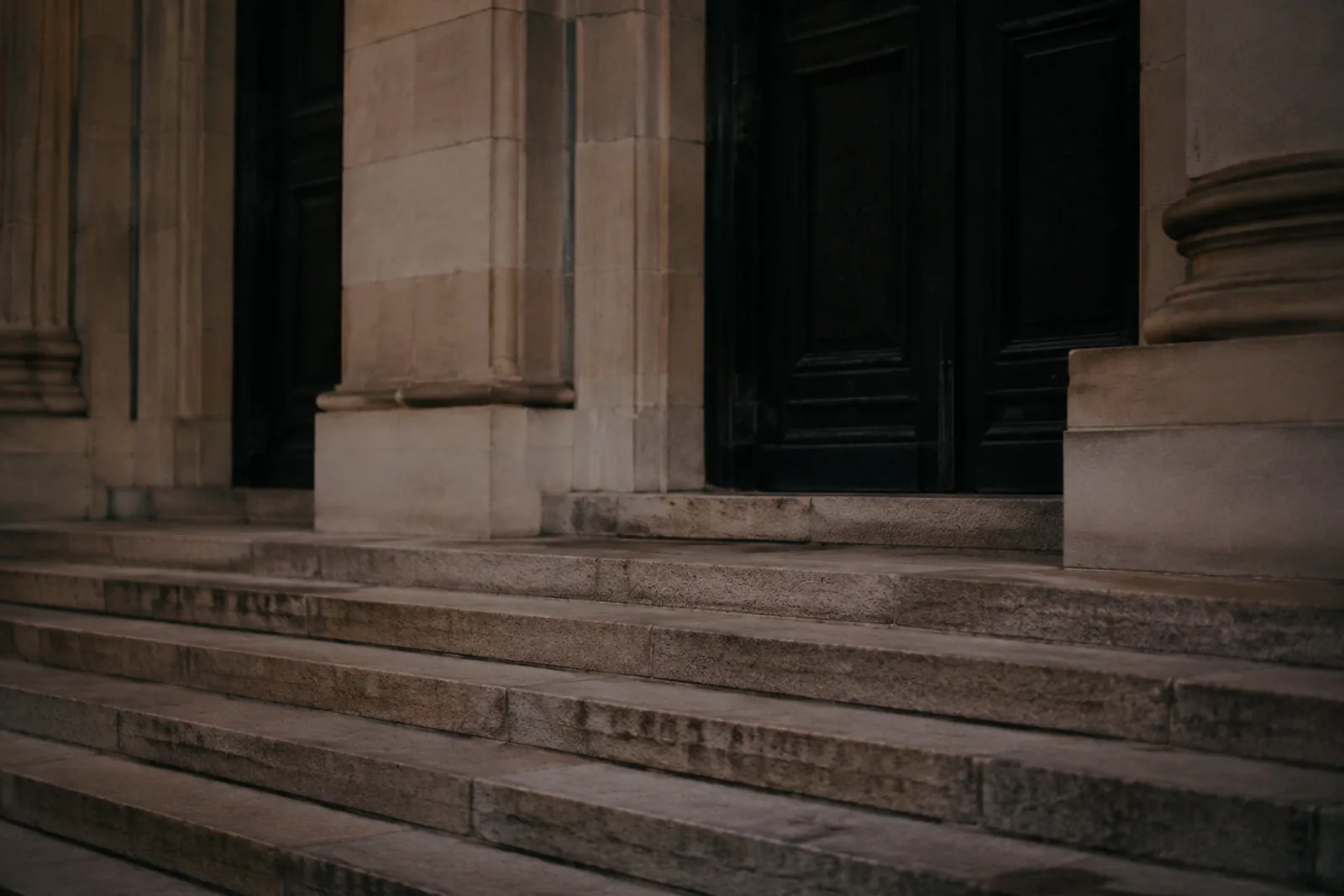 Courthouse stone steps at dusk, near-black doors and pale stone facade, close architectural composition excluding all lettering and symbols, no people.