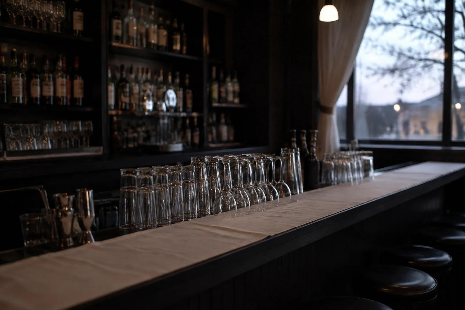 An independent neighborhood cocktail lounge before opening, dark wooden bar and simple empty glasses, white linen bar cloth, available dusk window light, no people.