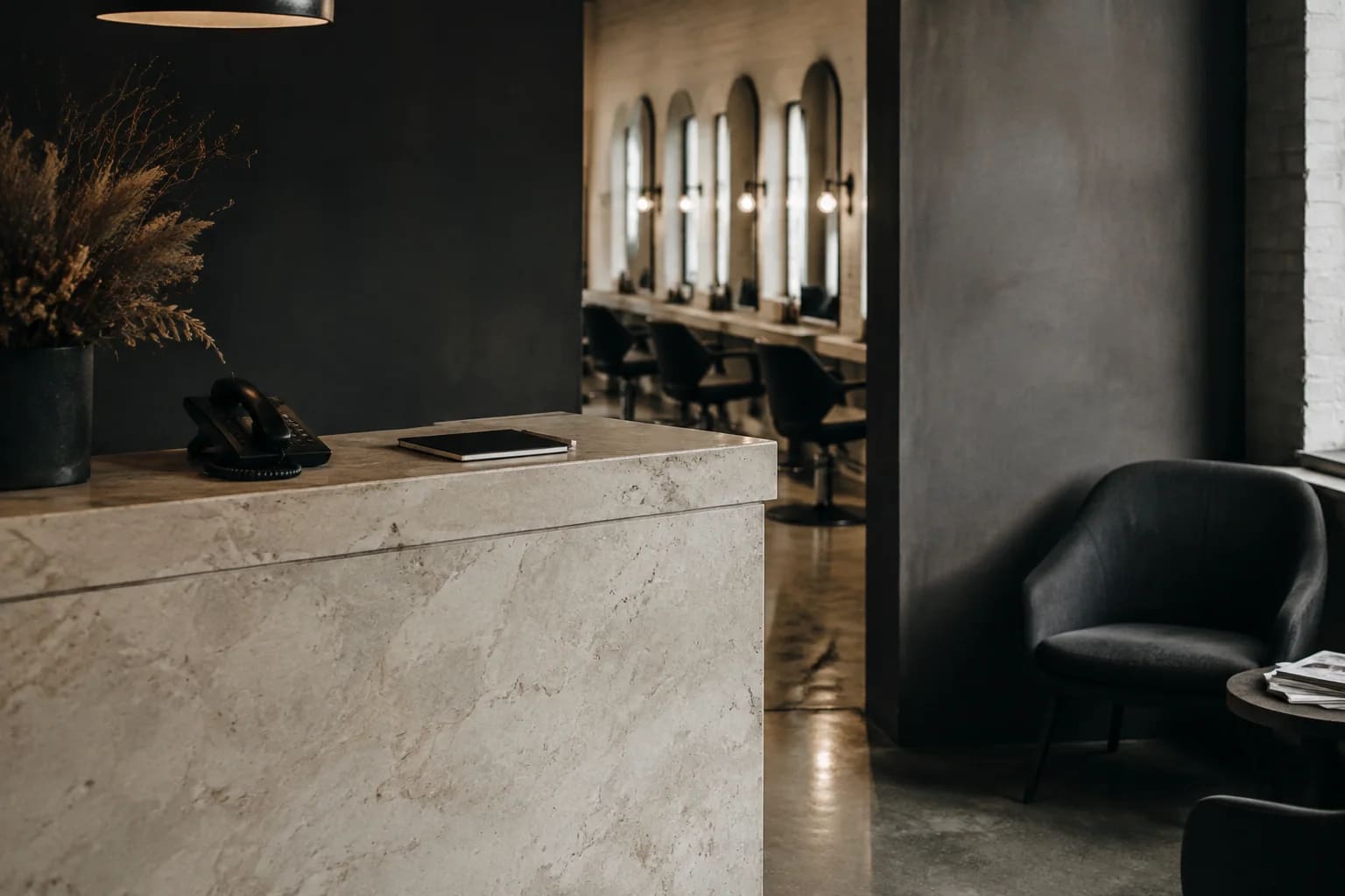 A small independent salon reception area with a pale stone desk, unbranded desk phone, closed appointment notebook, charcoal waiting chairs and a glimpse of mirrors beyond, natural window light.
