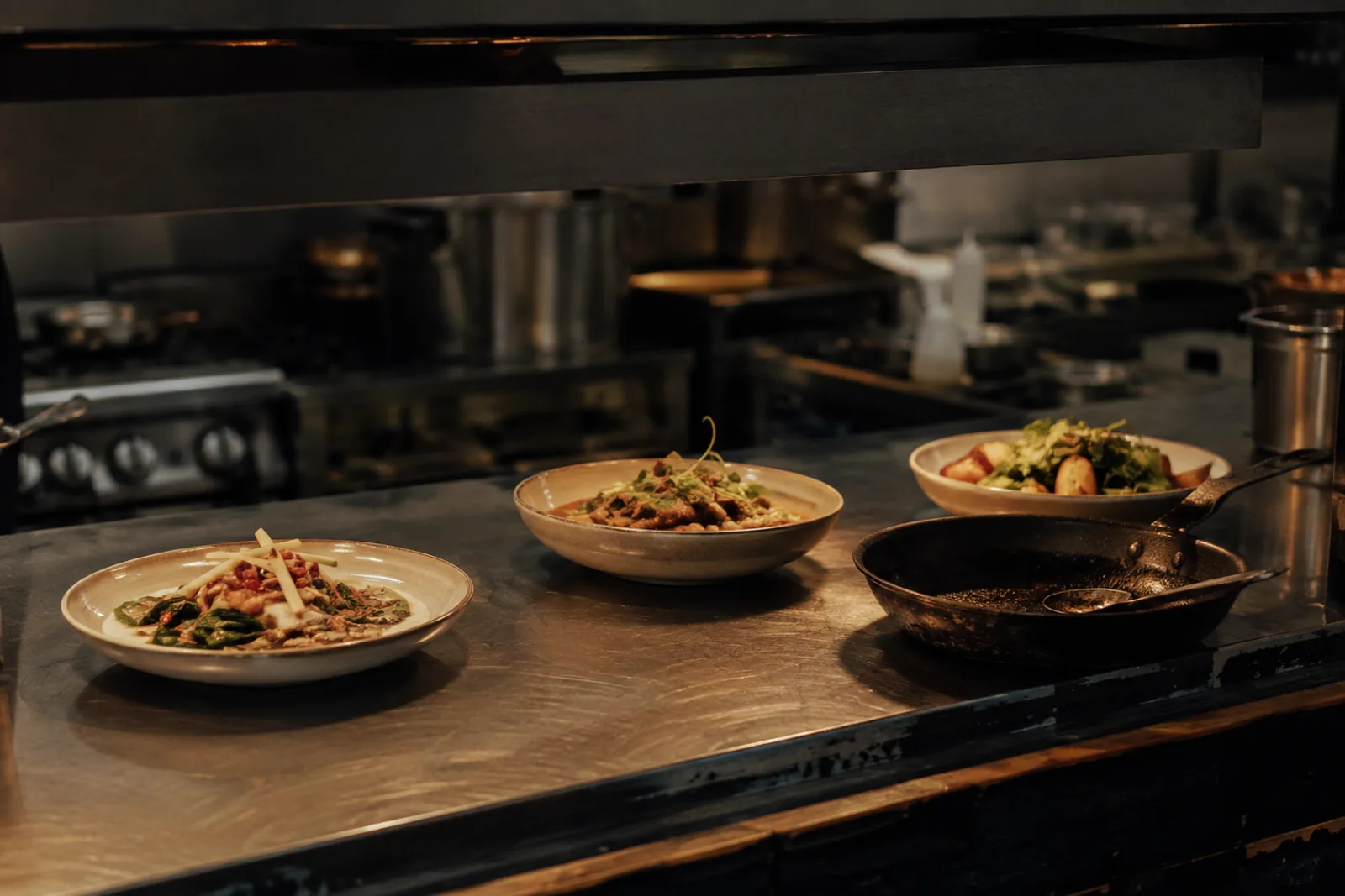 An independent restaurant pass window with three carefully plated dishes, stainless steel, a blackened pan, and warm practical light; an unposed moment before service, no people.