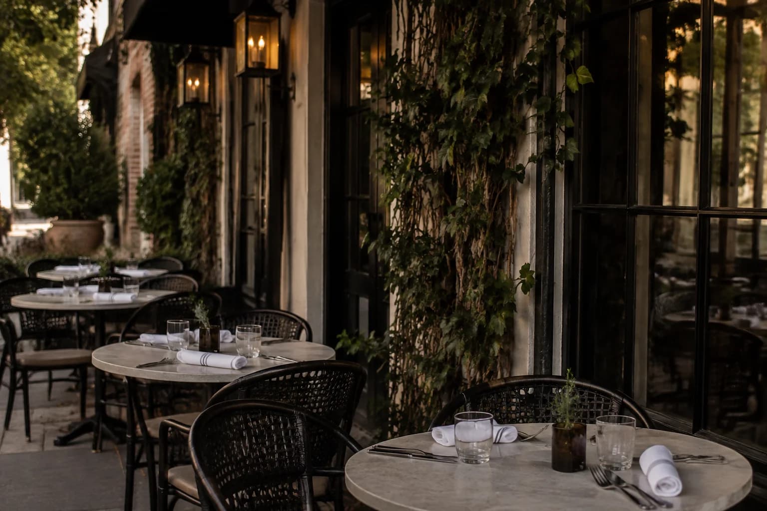 A neighborhood restaurant patio with dark bistro chairs, round stone tables, climbing vines and linen napkins, afternoon side light before service.